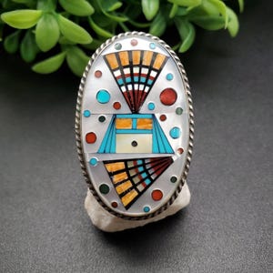 May include: Oval-shaped silver ring with a detailed inlay design. The ring features a central design with turquoise, orange, red, and white accents. The ring is set on a white stone, showcasing the intricate craftsmanship and vibrant colors.