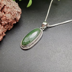 K&ouml;nnte beinhalten: Eine silberne Halskette mit einem gr&uuml;nen ovalen Anh&auml;nger. Der Anh&auml;nger ist in einer silbernen Fassung mit einem seilartigen Design gefasst.