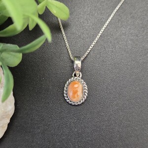 May include: A silver necklace featuring an oval pendant with a smooth, light orange stone set in a twisted rope-style metal border. The pendant hangs from a delicate silver box chain, showcasing a polished metallic finish and a simple design.