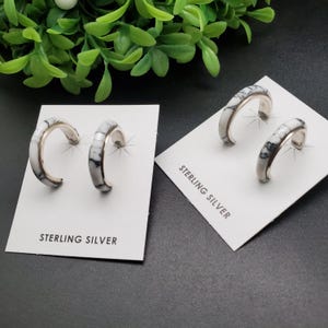May include: Two pairs of sterling silver hoop earrings with a white and black marbled design. Each pair is displayed on a white card with "STERLING SILVER" printed on it. Green foliage is in the background.