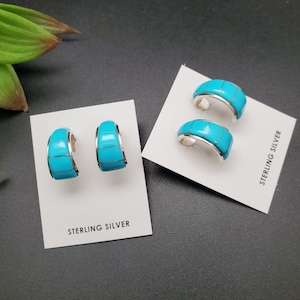 May include: Sterling silver earrings and a ring set with bright turquoise inlay. The jewelry is displayed on white cards against a dark background. The earrings are a hoop style, and the ring is a wide band. The text "STERLING SILVER" is printed on the cards.