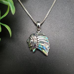 May include: Silver-toned pendant necklace featuring a Native American chief design. The pendant showcases a detailed profile and a headdress with iridescent abalone shell accents. The chain is a delicate link style, and the background is a dark, solid color.