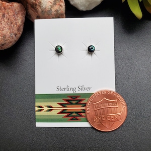 May include: A pair of sterling silver earrings with black abalone shell inlay. The earrings are displayed on a white card with the words "Sterling Silver" printed on it. A penny is included for size reference.