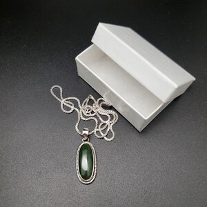 K&ouml;nnte beinhalten: Eine silberne Kettenkette mit einem gr&uuml;nen ovalen Anh&auml;nger. Der Anh&auml;nger ist in Silber gefasst und hat einen perlenbesetzten Rand. Die Kette befindet sich in einer wei&szlig;en Schachtel.