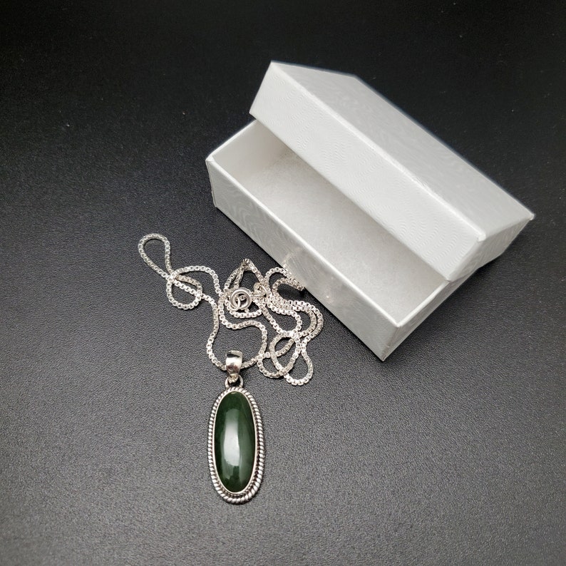 K&ouml;nnte beinhalten: Eine silberne Kettenkette mit einem gr&uuml;nen ovalen Anh&auml;nger. Der Anh&auml;nger ist in Silber gefasst und hat einen perlenbesetzten Rand. Die Kette befindet sich in einer wei&szlig;en Schachtel.