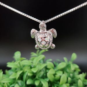 May include: A silver-colored turtle pendant necklace. The turtle pendant has a shell with an opal-like design and is encrusted with small, clear stones. The necklace chain is also silver-colored and has a box chain style.