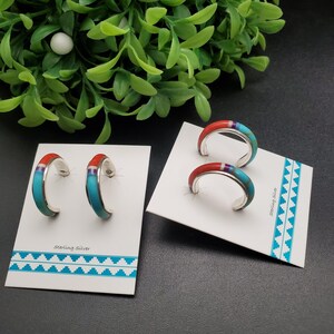 May include: Sterling silver hoop earrings with turquoise, red, and purple inlay. The earrings are displayed on white cards with a turquoise and white geometric pattern. The earrings have a polished finish and a curved design.