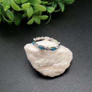 May include: A silver ring with alternating oval blue opal stones and small clear round stones. The ring is set on a white rock against a dark background, with green foliage in the upper left corner.