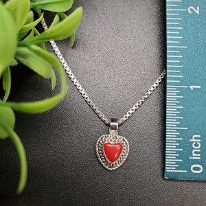 May include: A silver chain necklace with a red heart pendant. The pendant is set in a silver frame with a detailed design. The necklace is shown next to a ruler, indicating the size of the pendant.
