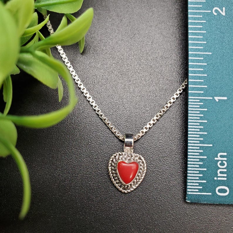 May include: A silver chain necklace with a red heart pendant. The pendant is set in a silver frame with a detailed design. The necklace is shown next to a ruler, indicating the size of the pendant.
