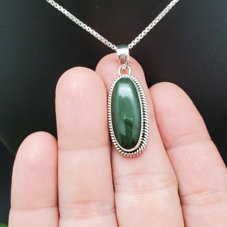 K&ouml;nnte beinhalten: Eine silberne Kettenkette mit einem ovalen Anh&auml;nger aus gr&uuml;nem Jade. Der Anh&auml;nger ist in einer silbernen Fassung mit einem seilartigen Rand gefasst.