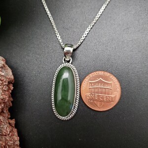K&ouml;nnte beinhalten: Eine silberne Kettenkette mit einem gr&uuml;nen Jade-Anh&auml;nger. Der Anh&auml;nger ist oval geformt und hat einen seilartigen Rand. Der Anh&auml;nger ist etwa so gro&szlig; wie ein deutscher Cent.