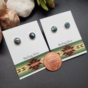 May include: Two pairs of sterling silver earrings with abalone shell inlays. The earrings are displayed on white cards with a green and brown patterned border. The text "Sterling Silver" is printed on the cards. A penny is included for scale.