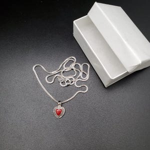 May include: A silver chain necklace with a heart-shaped pendant. The pendant has a red stone in the center.
