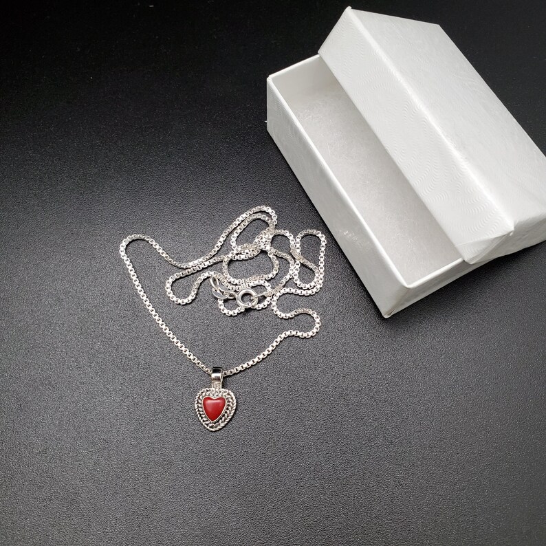 May include: A silver chain necklace with a heart-shaped pendant. The pendant has a red stone in the center.