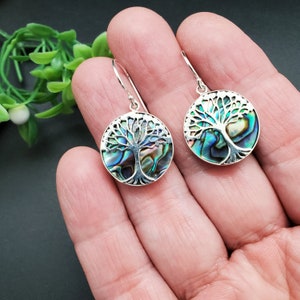 May include: A pair of silver earrings with a tree of life design. The earrings are made with abalone shell and have a round shape.