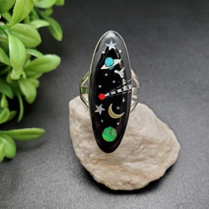 May include: A silver ring featuring a black oval stone set with colourful inlays depicting a space scene with planets, stars, and a comet.