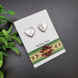 May include: A pair of sterling silver heart-shaped earrings with white mother-of-pearl inlays. The earrings are displayed on a white card with a green, red, and yellow patterned border. The card reads "Sterling Silver".