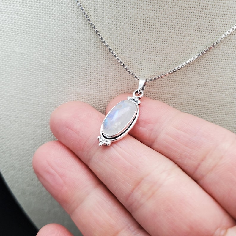 May include: A silver chain necklace with a white oval moonstone pendant. The pendant is set in a silver frame with decorative details.