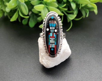 Könnte beinhalten: Ein silberner Ring mit einem ovalen Stein, der in der Mitte eingesetzt ist. Der Stein ist mit Türkis, Rot, Schwarz und Weiß in einem geometrischen Muster eingelegt. Der Ring liegt auf einem weißen Stein.