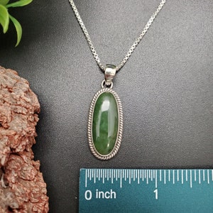 K&ouml;nnte beinhalten: Eine silberne Kettenkette mit einem gr&uuml;nen ovalen Anh&auml;nger. Der Anh&auml;nger ist in einen silbernen Rahmen mit einem seilartigen Design gefasst. Der Anh&auml;nger ist etwa 2,5 cm lang.