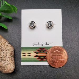 May include: A pair of sterling silver earrings with a light blue gemstone in the center of each earring. The earrings are displayed on a white card with the words "Sterling Silver" printed on it. A penny is included for size reference.