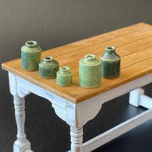 May include: Miniature green ceramic jars of different sizes are displayed on a small wooden table with white distressed legs. The jars have a glossy finish and are in shades of green.