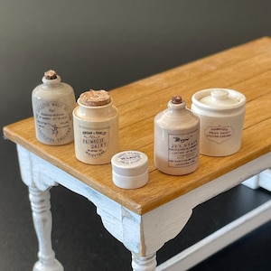 May include: A collection of miniature antique-style jars and bottles with labels, displayed on a small wooden table with white distressed legs. The collection includes a small jar with a cork stopper and a small white jar with a lid.