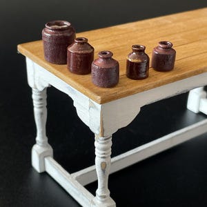 May include: A collection of miniature ceramic jars in shades of burgundy, displayed on a small wooden table with white, distressed legs. The jars have a textured finish. The table is light brown. The table's height is approximately 70 cm.