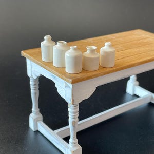 May include: A miniature wooden table with a white-painted base and a light brown tabletop. Six small, off-white ceramic bottles are arranged on the table. The table has a distressed, vintage appearance, with visible wear on the paint.