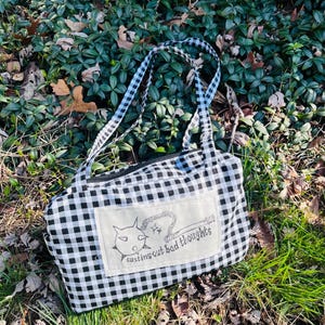 May include: A black and white checkered tote bag with a zipper closure and matching handles. The bag features a rectangular patch with the text "casting out bad thoughts" and a cartoon illustration. The bag is sitting on a bed of leaves and grass.