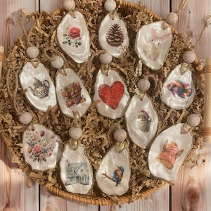 May include: Twelve unique Christmas ornaments made from oyster shells with various designs, including a heart, a house, a bird, a pine cone, a teapot, a Santa Claus, an angel, and a floral pattern. The ornaments are displayed in a wicker basket filled with brown packing paper.