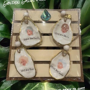 Op de afbeelding: Een set van vier Golden Girls-thema ornamenten. Elk ornament heeft de vorm van een oesterschelp met een gouden rand, met het gezicht van een personage en een citaat: "Think like Dorothy", "Love like Blanche", "Live like Rose" en "Speak like Sophia".