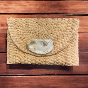 May include: A woven straw clutch with a natural oyster shell clasp. The clutch is a light beige color and has a rectangular shape.