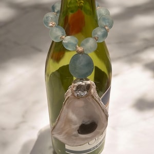 May include: A green wine bottle adorned with a necklace of light blue glass beads and a large, matching pendant. An oyster shell bottle stopper is attached to the bottle, adding a unique, coastal touch to the design.
