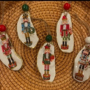 May include: Five Christmas ornaments shaped like oyster shells with painted nutcracker figures. Each ornament features a different nutcracker design with varying colors and details. The ornaments are decorated with gold accents and have a rustic, vintage aesthetic.