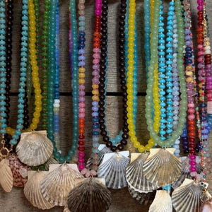May include: A collection of necklaces featuring strands of colorful glass beads in shades of blue, yellow, green, pink, and black, each accented with a natural seashell pendant hanging from a metal clasp attached to a display rack.