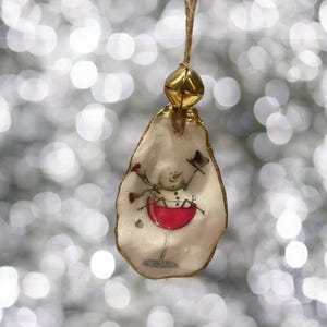 May include: A white oyster shell ornament with a hand-painted snowman holding a glass of red wine. The ornament has a gold trim and a gold bell at the top.