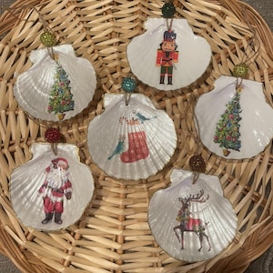 May include: Six white seashell ornaments with gold trim, each featuring a different Christmas-themed image: a Christmas tree, a stocking with candy canes, a Santa Claus, a reindeer, a nutcracker, and another Christmas tree.