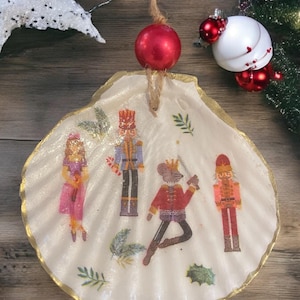 May include: A white seashell ornament with a gold trim, featuring a Nutcracker theme. The ornament depicts a Nutcracker soldier, a mouse king, a ballerina, and a candy cane. The ornament is decorated with green leaves and holly.