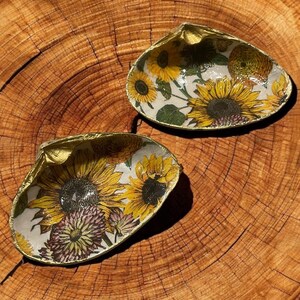 May include: Two golden seashells with a white background and a sunflower and floral pattern. The shells are on a wooden surface.