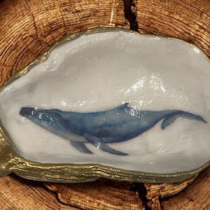May include: A decorative oyster shell dish with a hand-painted blue whale design. The interior is white, and the rim is trimmed with gold. The dish is set on a wooden surface.