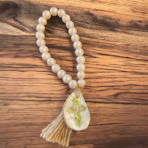 May include: A white beaded garland with a tassel and a white seashell with a green floral cross design.
