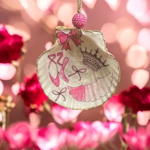 May include: A white seashell ornament with a pink ballerina design. The ornament features a crown, a tutu, ballet shoes, and bows. The seashell is decorated with gold paint around the edges.