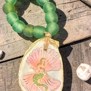 May include: A handmade seashell pendant featuring a mermaid illustration, with a gold-painted edge, and a strand of green glass beads. The mermaid has pink wings and a green tail. The pendant is attached to the beads with twine.