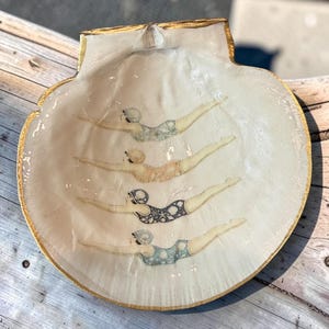 May include: A large, off-white scallop shell dish with a gold-painted rim. The interior features four illustrations of swimmers in vintage-style swimsuits, each wearing a swim cap and positioned in a horizontal diving pose, arranged in a vertical row.