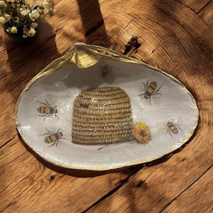 May include: A white ceramic dish with a gold rim, featuring a beehive and bees. The dish is decorated with a sunflower and has a gold leaf design.