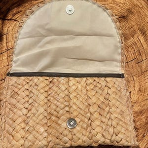 May include: A woven straw clutch bag with a brown leather trim and a snap closure. The bag is lined with a beige fabric.
