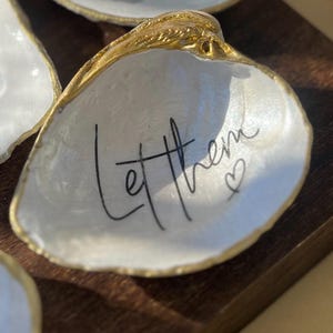 May include: A white seashell dish with gold trim and the words "Let them" written in black calligraphy. A small heart is drawn next to the text.