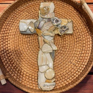 May include: A decorative cross made of various seashells in shades of white, beige, and gold. The cross is placed on a woven wicker tray. The seashells are arranged to create a mosaic-like pattern, adding texture and visual interest.